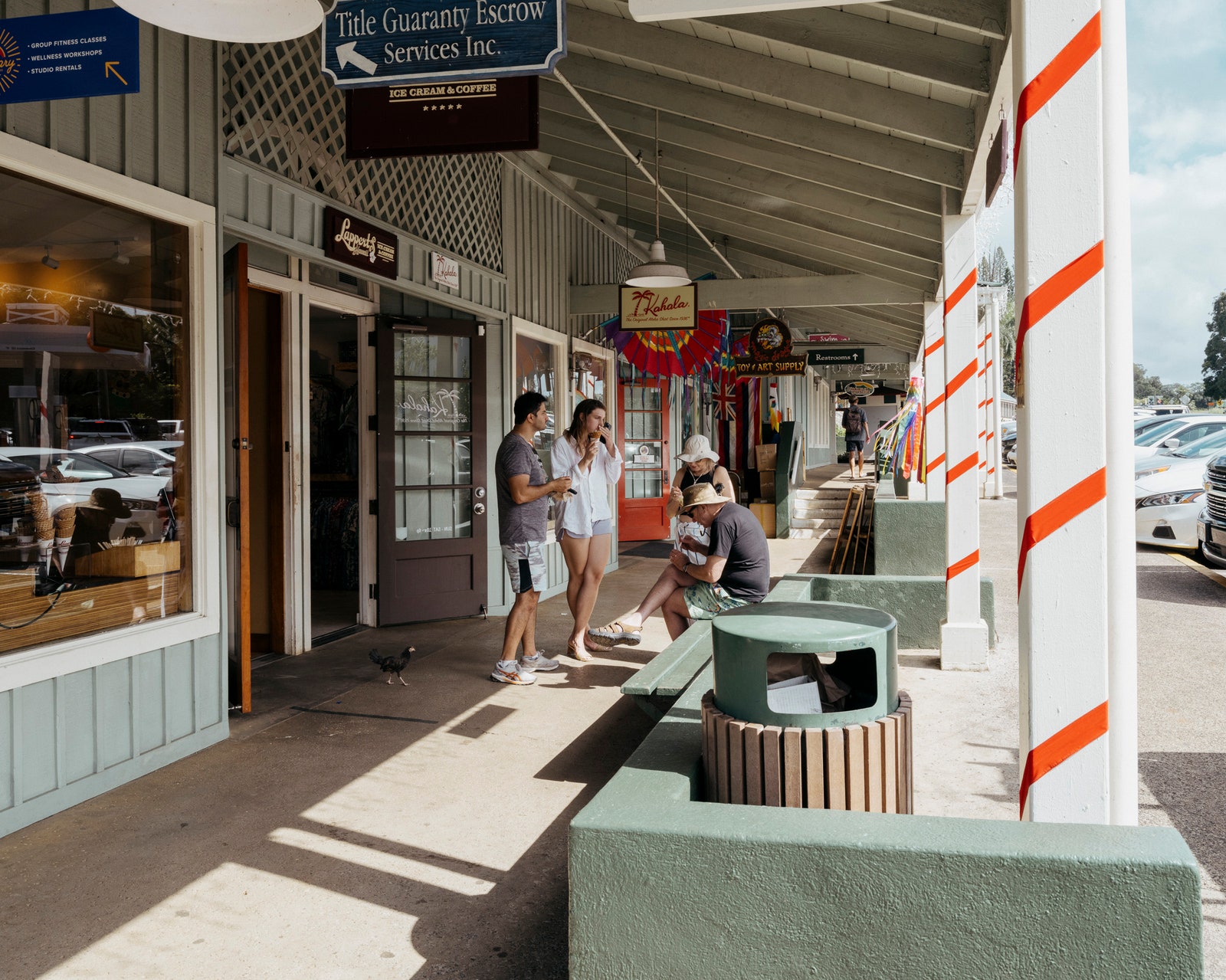 A small strip mall with people eating ice cream outside as a chicken walks by