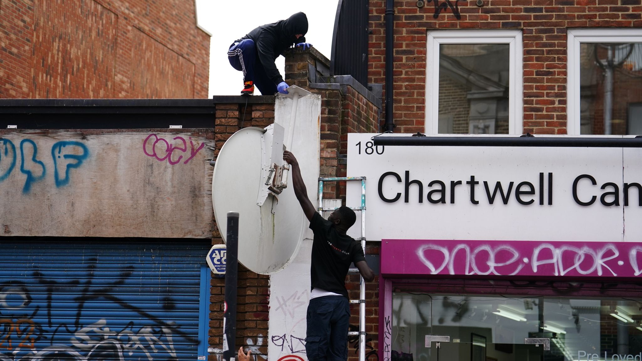 People remove a new artwork by Banksy, depicting a howling wolf painted on a satellite dish that was placed on a shop roof in Peckham, south London. The artist's latest artwork comes a day after he unveiled three monkeys painted on a bridge in Brick Lane, east London. Picture date: Thursday August 8, 2024. 