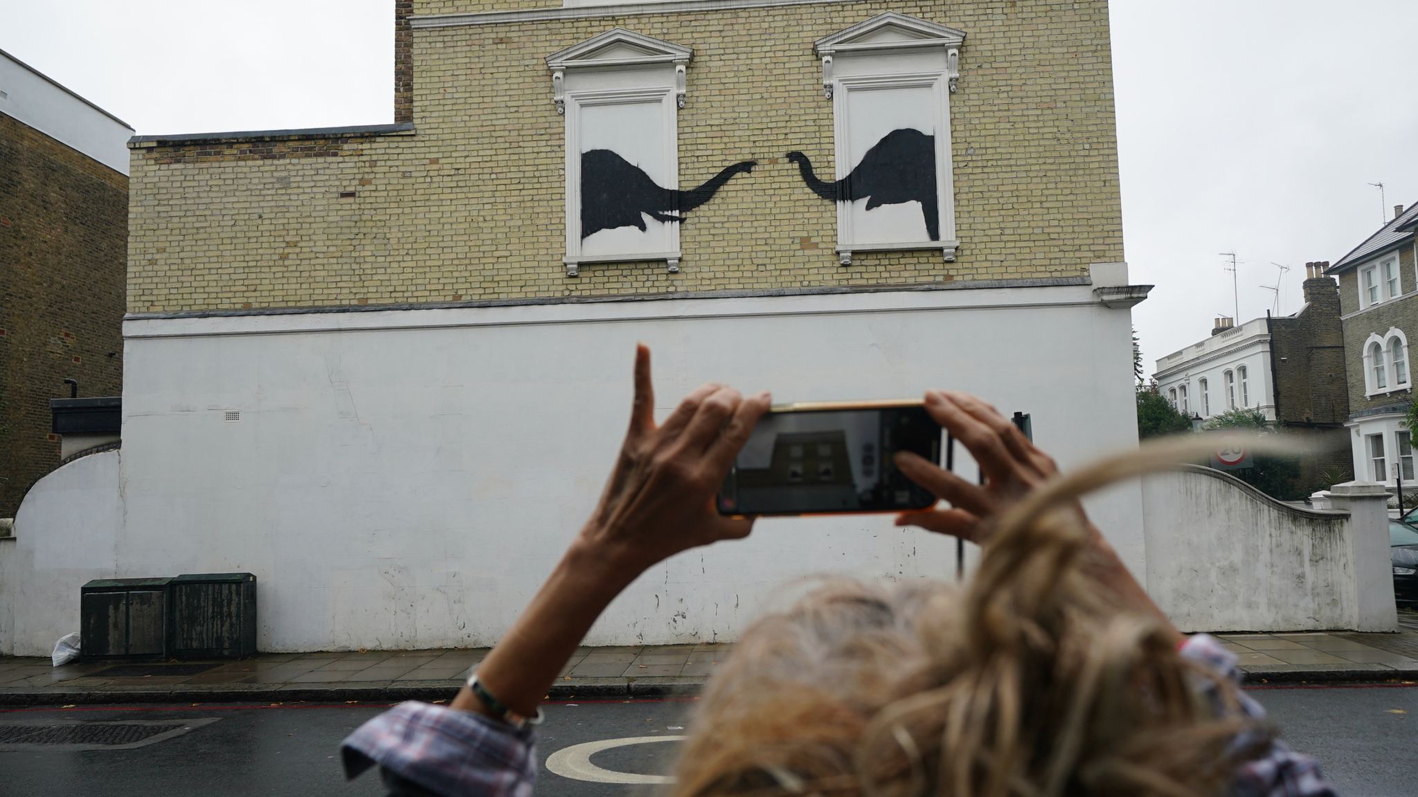 A woman stops to photograph a new artwork depicting two elephants poking their heads out of blocked out windows unveiled by Banksy on the side of a building at the junction of Edith Grove and Edith Terrace, in Chelsea, south west London. Picture date: Tuesday August 6, 2024.